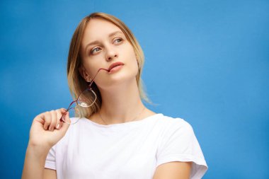 Beyaz tişörtlü çekici genç bir sarışın hayallere dalmış gibi görünüyor, gözlüğünü ağzında tutuyor ve yüzünde ciddi bir ifade var..