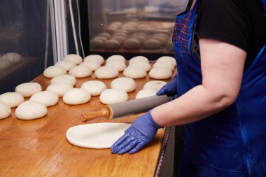 Önlüklü ve mavi eldivenli yetişkin bir dişi fırıncı, hamuru elleri ve mutfak masasında bir oklavayla yuvarlıyor. Pasta pişirme, pide, kek, kurabiye, fırın ürünleri yapma, ekmek pişirme kavramı.