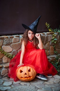 9 yaşında, kırmızı elbiseli bir kız dışarıda bir cadılar bayramı partisinde çitin yanında, parkta bir balkabağıyla oturuyor. Muhteşem bir sonbahar bayramı konsepti