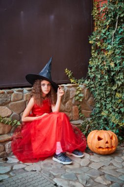 9 yaşında, kırmızı elbiseli bir kız dışarıda bir cadılar bayramı partisinde çitin yanında, parkta bir balkabağıyla oturuyor. Muhteşem bir sonbahar bayramı konsepti