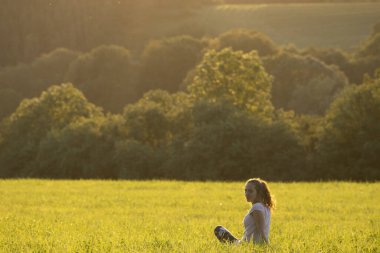 Genç kız güneşli bir çayırda oturuyor ve gülümsüyor.