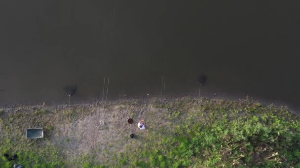 ドローンで撃つ トップビュー 池の岸にある白いTシャツを着たひげを生やした漁師が 魚を釣る準備をしている — ストック動画