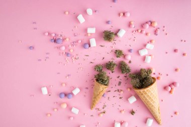 Kuru tıbbi marihuana tomurcukları pembe arka plandaki kremalı dondurma külahlarının üzerinde yatıyor. Etrafta şekerler ve marşmelovlar var. Alternatif tıbbi esrar tedavisi