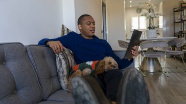 Tablet kullanan bir adam, köpek kucağında dinleniyor.