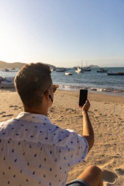 Plajda akıllı telefon kullanan kulaklık takan bir adam.