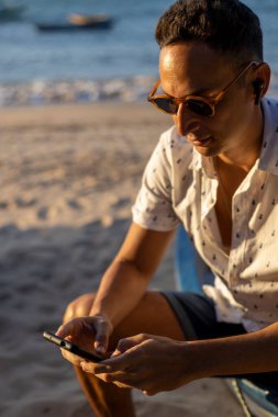 Sahilde oturan ve akıllı telefon kullanan bir adam.