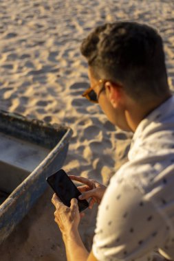 Sahilde oturan ve akıllı telefon kullanan bir adam.