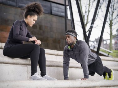 Athletic woman assisting man doing push-ups on steps outdoors