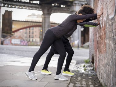 Athletic man and woman stretching against wall outdoors