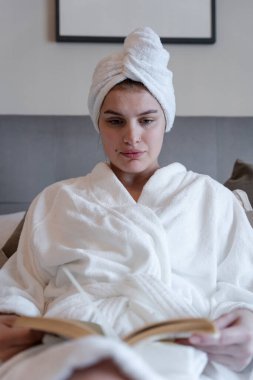 Bornozlu kadın yatakta kitap okuyor.