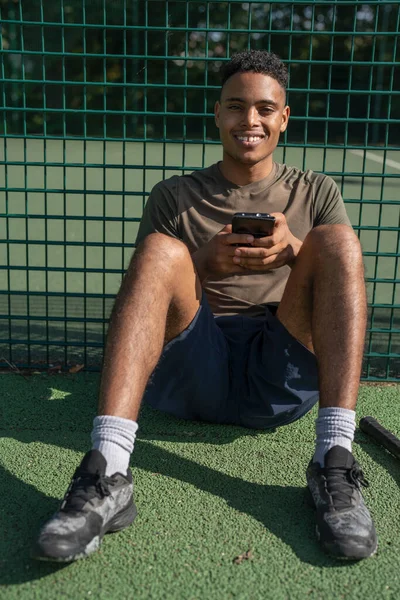 Akıllı telefonuyla tenis kortunda oturan gülümseyen bir adamın portresi.