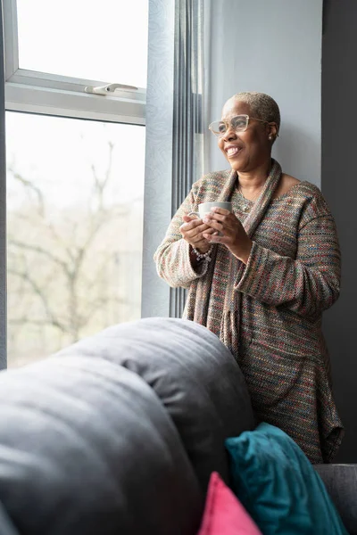 Elinde kahve bardağıyla pencereden bakan olgun bir kadın.