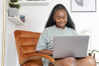 Evde koltukta otururken dizüstü bilgisayar kullanan genç kadın