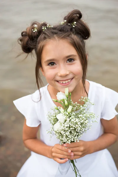 Una bambina tiene un mazzo di fiori bianchi su uno sfondo sfocato.capelli scuri, fiori bianchi nelle sue mani, vestito bianco — Foto Stock