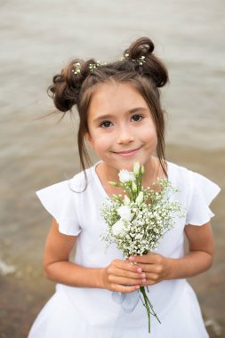 Bir kız çocuğu bulanık bir arkaplanda bir buket beyaz çiçek tutar. Siyah saçlar, ellerinde beyaz çiçekler, beyaz elbiseler.