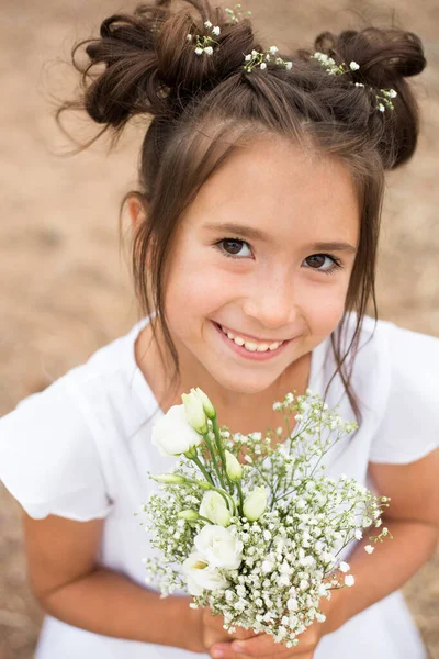 Une enfant fille tient un bouquet de fleurs blanches sur un fond flou.cheveux foncés, fleurs blanches dans ses mains, robe blanche — Photo