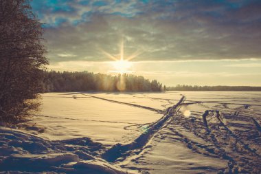 Karla kaplı bir göl üzerinde güneşli bir gün batımı. Bulutlu mavi gökyüzü. Kar üzerinde kayak izleri ve patikalar.