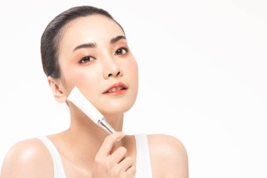 Güzel, Asyalı, kahverengi saçlı, gülümseyen bir kadın. Mutluluktan gülümsüyor ve elinde makyaj fırçasıyla kozmetik krem losyonu ve neşeli bir şişe tutuyor. Beyaz arka planda güzellik kavramı..