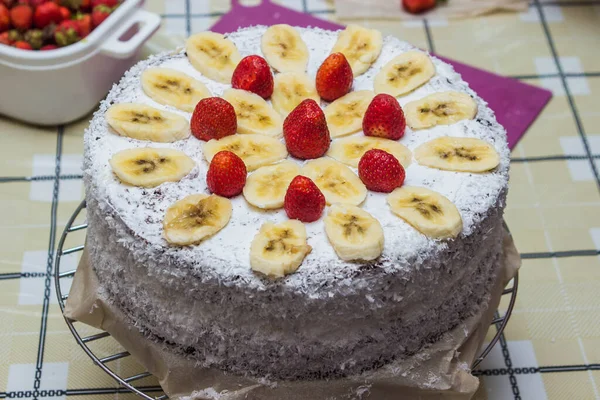Homemade fruit cake with cream. Cake decorated with strawberries and bananas. Sprinkled with coconut on the sides. Photo of cake in the home kitchen
