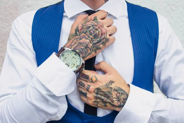 The guy straightens his tie. Hands in tattoos. A guy in a shirt and a blue jacket. watch on his right hand