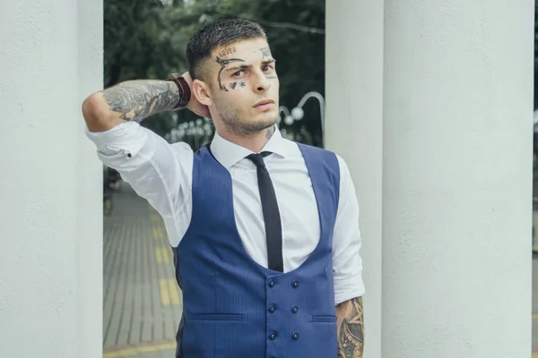 The guy holds his hand behind his head. The guy in tattoos. A guy in a suit in the park. Stylish and elegant look