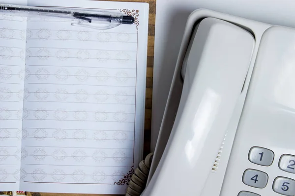 A blank sheet of notebook lies next to a landline telephone. Top is a ballpoint pen. The concept of business calls. Cold calls.