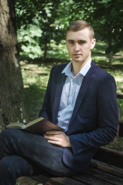 Adam bir kitap okuyor. Takım elbiseli genç bir adam parkta bir bankta otururken kitap okuyor.