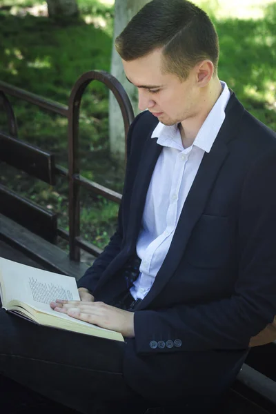 The guy is reading a book. A young guy in a suit reads a book while sitting on a bench in a park