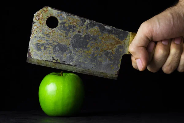 Green apple on a black background. Hand holds a kitchen hatchet over an apple. Hatchet old with rust