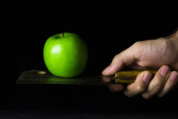 Green apple on a black background. An apple is standing on a kitchen hatchet. Hatchet old with rust