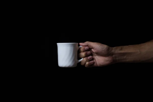 A man\'s hand holds a white porcelain mug on a black background. Place for an inscription or logo on the mug