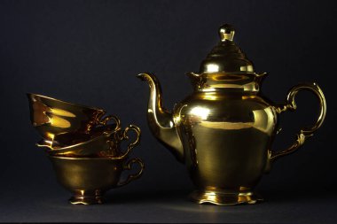 Siyah arka planda altın çay seti. Çay fincanları üst üste yığılmış ve yakınlarda bir çaydanlık var.