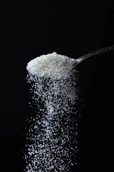 sugar is poured from a spoon on a black background. Excessive sugar intake. Sweet crystals