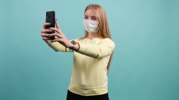 Chica joven con máscara médica en la cara saluda a los amigos durante la conversación en línea. — Vídeos de Stock