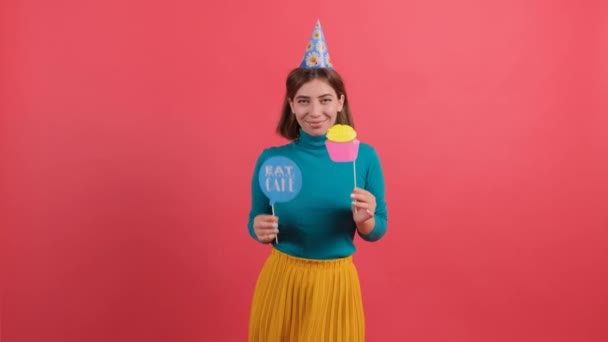 Joyeux jeune femme en chapeau de fête tenant accessoires d'anniversaire sur fond rouge. — Video