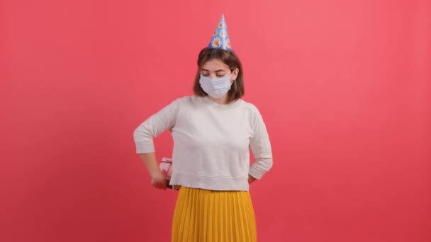 Оптимістична дівчина з медичною маскою на обличчі і конусом на голові дарує подарункову коробку — стокове відео