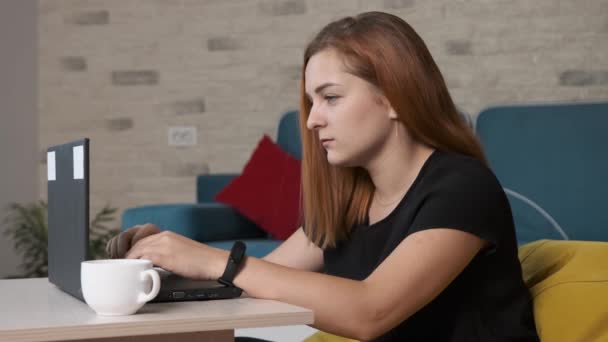 Mujer joven está bebiendo una taza de café mientras está trabajando en el ordenador portátil de la oficina en casa. — Vídeos de Stock
