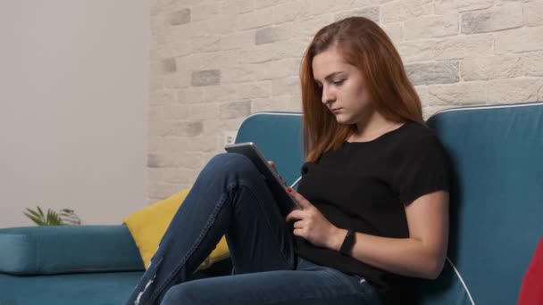 Serieus meisje zit op een bank en werkt geconcentreerd op de tablet. — Stockvideo