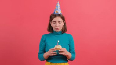Doğum günü şapkalı genç kadın pastaya mum üflüyor, kırmızı arka planda izole edilmiş..