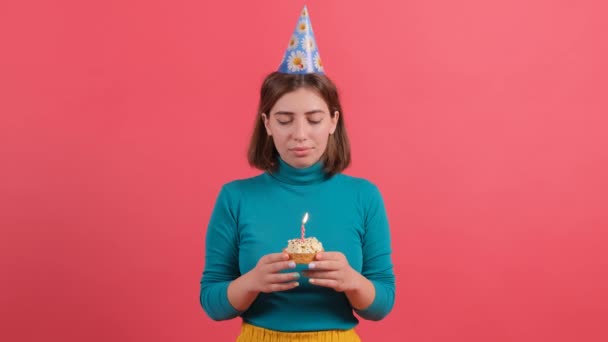 Молода жінка в капелюсі на день народження вибухає свічкою на торт, ізольована на червоному тлі . — стокове відео