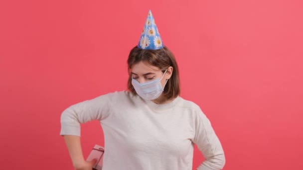 Optimistisches Mädchen mit medizinischer Maske im Gesicht und Kegel auf dem Kopf, die Geschenkbox schenkt. — Stockvideo