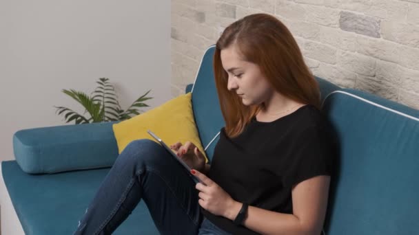 Chica seria está sentado en un sofá y trabajando concentrado en la tableta. — Vídeos de Stock