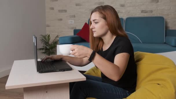 Jeune femme boit une tasse de café tout en travaillant à l'ordinateur portable à partir du bureau à la maison. — Video