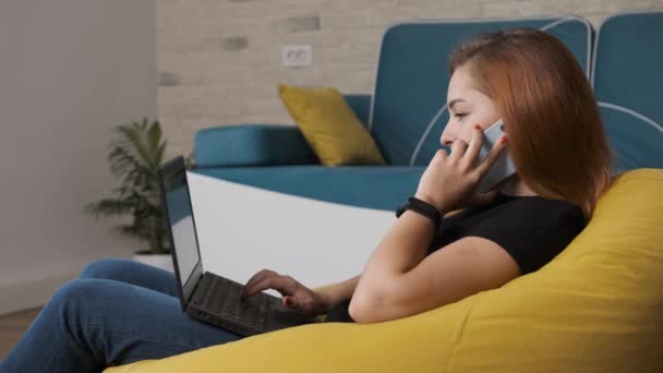 Jovem mulher está trabalhando no laptop enquanto está falando no telefone. — Vídeo de Stock