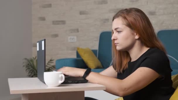 Jovem mulher está bebendo uma xícara de café enquanto está trabalhando no laptop do escritório em casa. — Vídeo de Stock