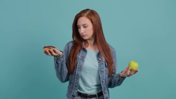 Молодая женщина выбирает между яблоком и кексом, девушка путается в своем выборе — стоковое видео
