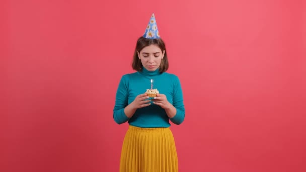 Jonge vrouw in verjaardag hoed blazen kaars op taart, geïsoleerd op rode achtergrond. — Stockvideo