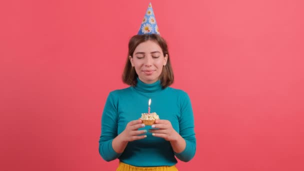 Молода жінка в капелюсі на день народження вибухає свічкою на торт, ізольована на червоному тлі . — стокове відео