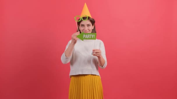 Parti şapkalı mutlu genç kadın kırmızı arka planda doğum günü aksesuarları tutuyor.. — Stok video