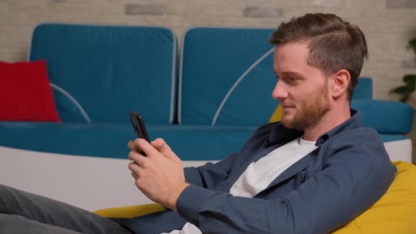 Giovane uomo si siede sul sacchetto di fagioli e giochi al telefono, a casa. — Video Stock